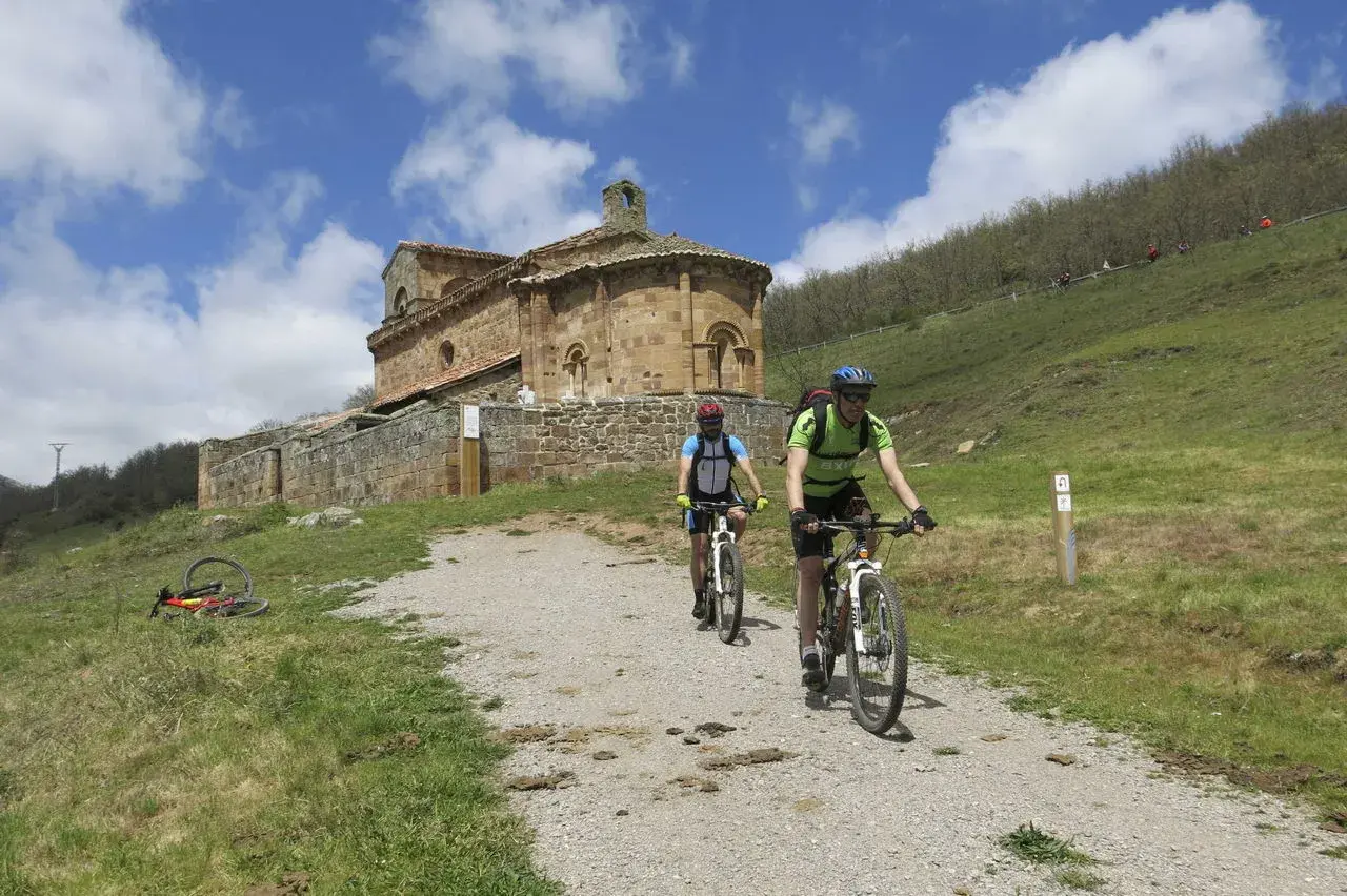 Ciclistas exploran el románico palentino, pedaleando hacia una iglesia de piedra bajo un cielo azul con nubes.
