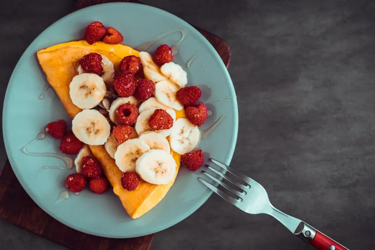 Omlet z bananem i malinami, polany syropem, na jasnoniebieskim talerzu. Obok leży widelec.