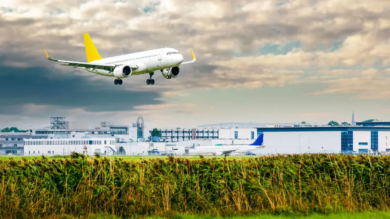 Ein Linienflugzeug mit gelbem Leitwerk nähert sich dem Flughafen. Die Linienflug Geschwindigkeit ist beeindruckend, während es über das Schilf und die Gebäude fliegt.