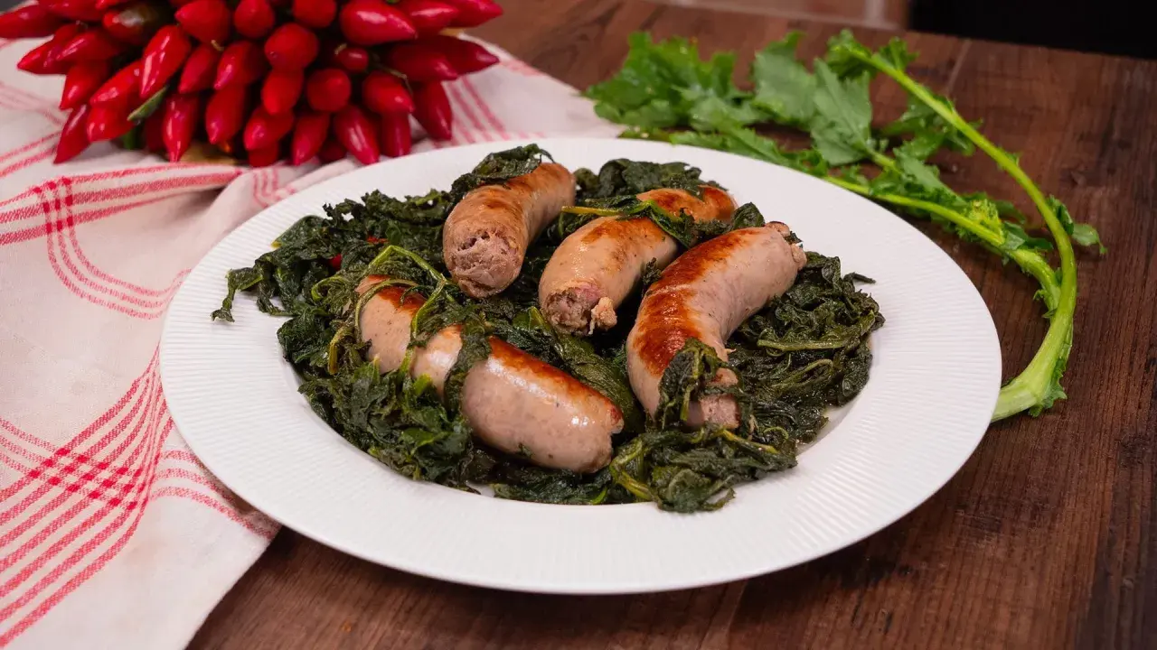 Salsiccia e friarielli, un piatto tradizionale napoletano, servito su un piatto bianco con peperoncini rossi sullo sfondo.
