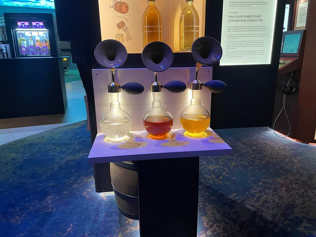 Tre flaskor med olika färger av sprit står upplysta på en hylla. En utställning på Vin & Sprit Museum.