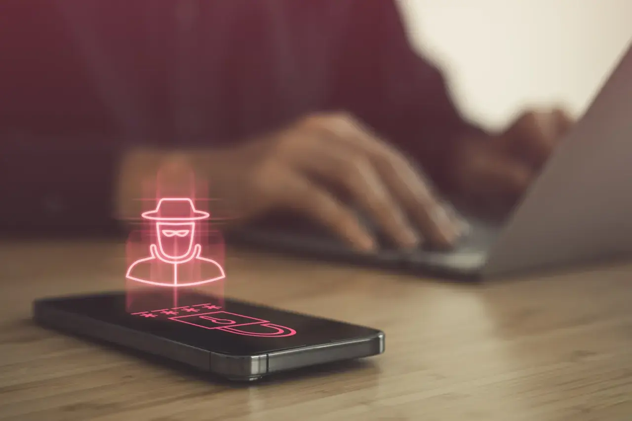 Haker w kapeluszu i gwiezdki symbolizujące hasło na ekranie telefonu, sugerujące podsłuch w telefonie. W tle osoba pracuje na laptopie.