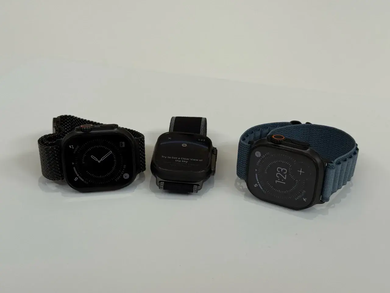 Trzy zegarki Apple Watch na białym tle: jeden z czarną bransoletą, drugi z czarną, trzeci z niebieską.