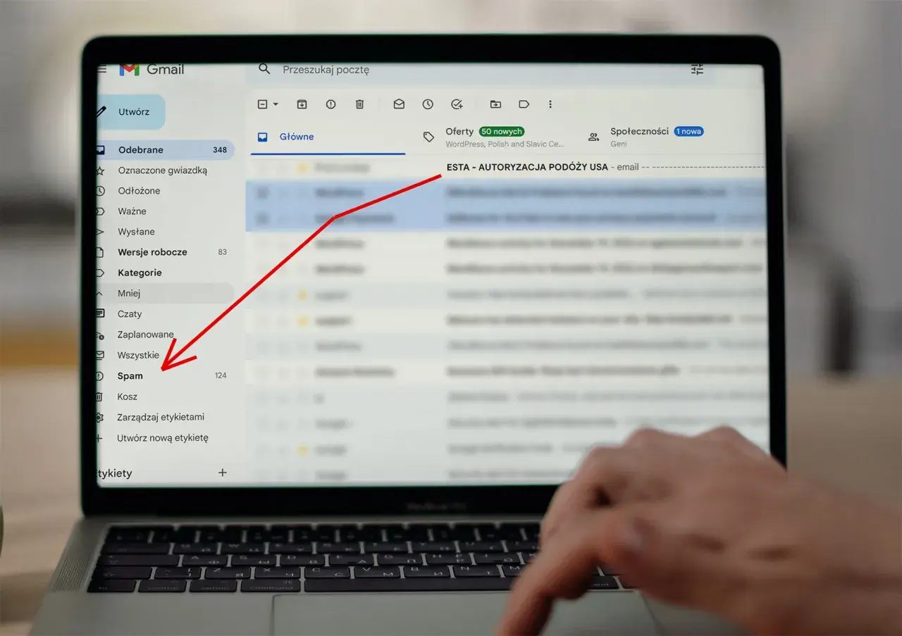 Laptop z otwartym mailem Gmail, zaznaczony folder "Spam" i czerwona strzałka wskazująca na temat maila "ESTA - AUTORYZACJA PODÓŻY USA".