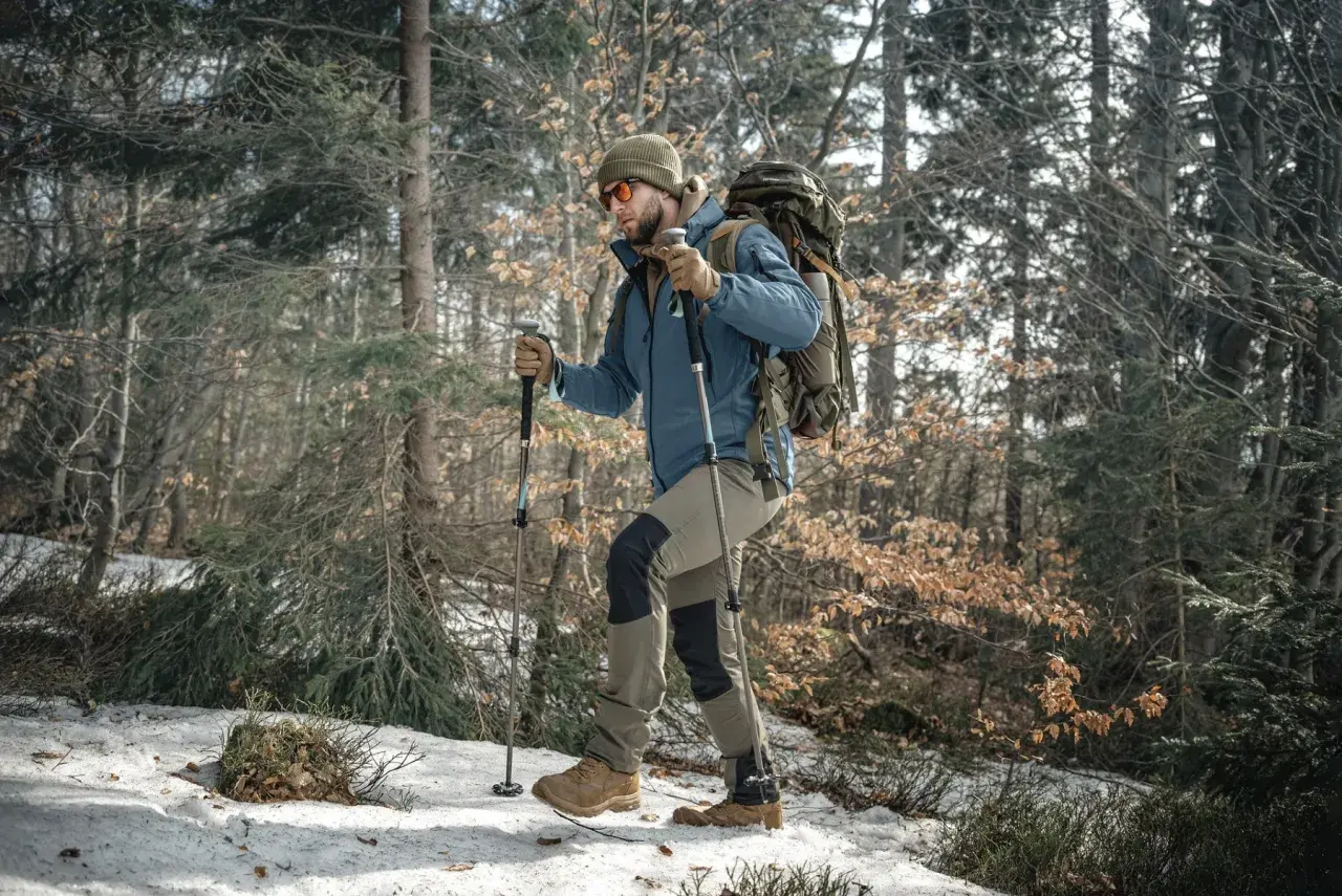 Mężczyzna w zimowej kurtce i czapce idzie przez las, wspierając się na kijach trekkingowych. W tle drzewa i śnieg.