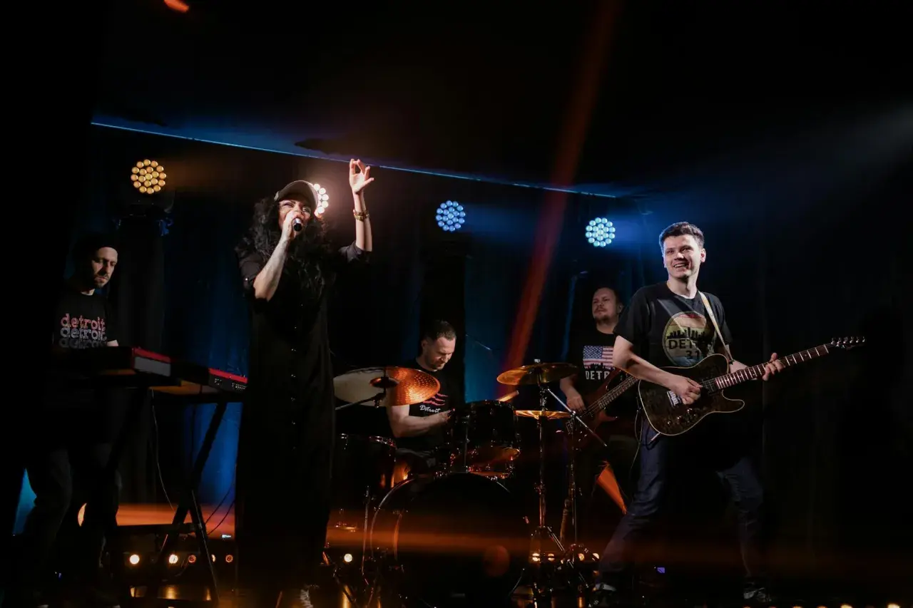 Energiczny zz trio rockowe na scenie. Wokalistka w czarnej sukience, gitarzysta, basista, perkusista i klawiszowiec tworzą dynamiczny zespół.