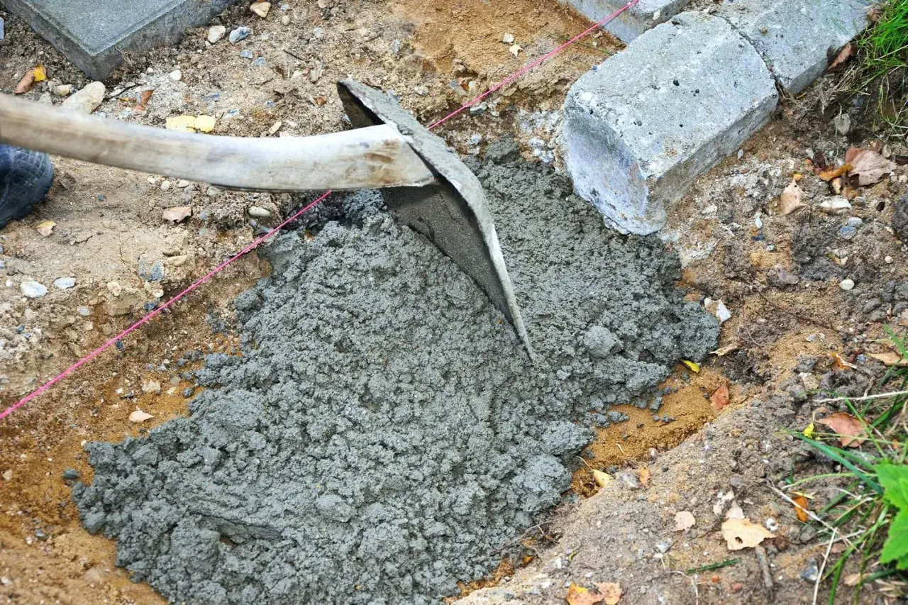 En hacka sprider jordfuktig betong i en grop. Betongblock ligger bredvid.