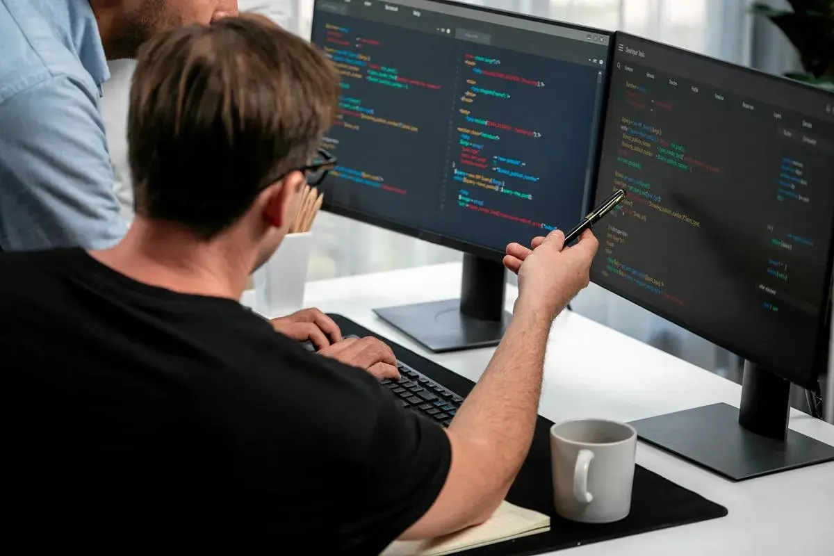 Dwóch programistów analizuje kod na monitorach. To świetny przykład, jak wygląda praca w IT dla początkujących – nauka i współpraca.