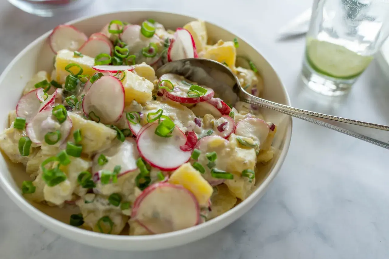 En krämig grekisk potatissallad med rädisor och gräslök, serverad i en vit skål med en sked.