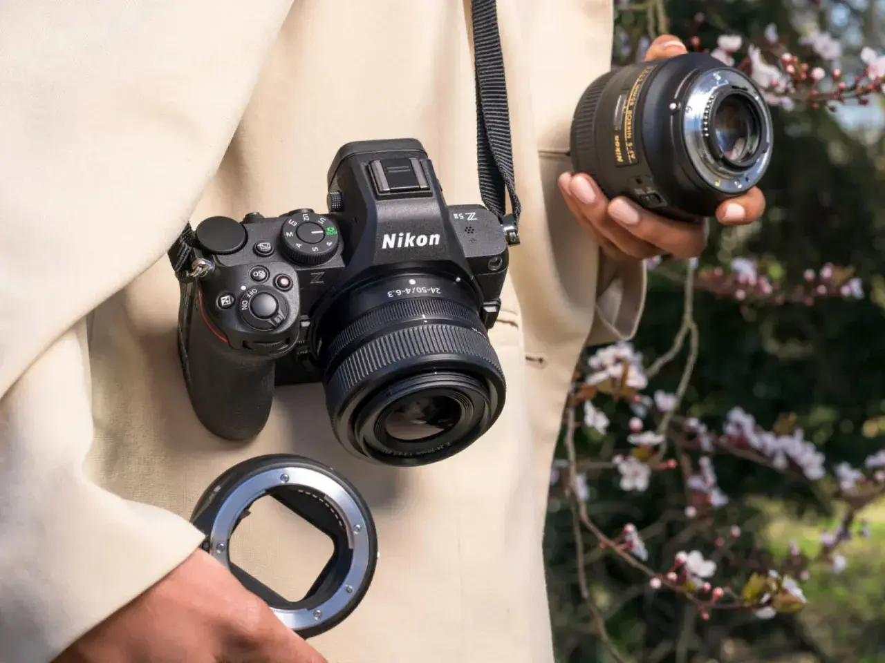 Osoba trzyma aparat Nikon Z5II z obiektywem i adapterem. W tle kwitnące drzewa.