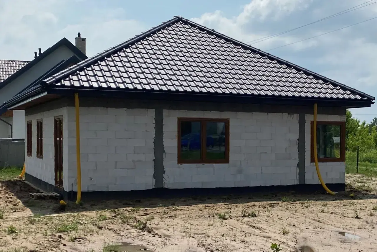 Dom w budowie z żółtymi rurami odprowadzającymi wodę z rynien. Widać białe bloczki, brązowe okna i dach pokryty ciemną dachówką.