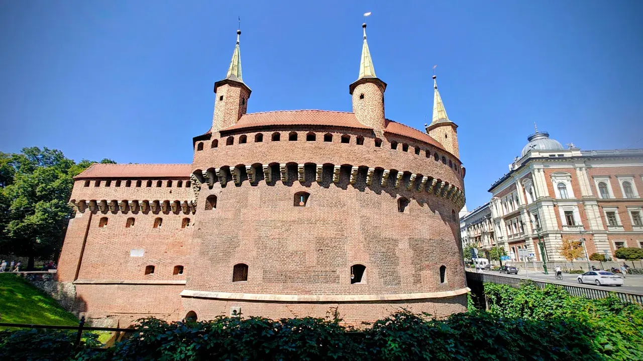 Krakowski zabytek przy Bramie Floriańskiej, okrągła baszta obronna z cegły, góruje nad zielonymi terenami i ulicą.