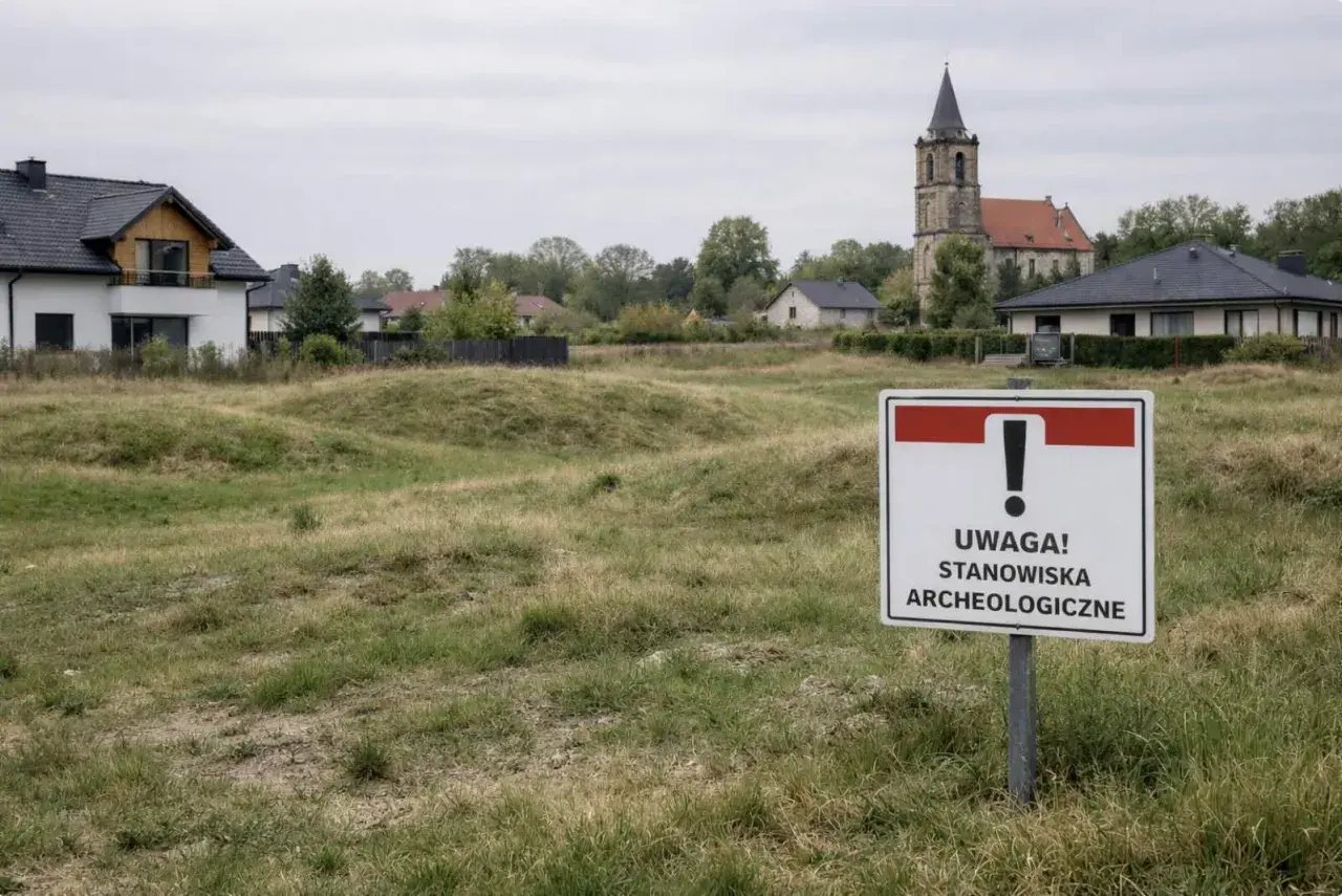 Znak "Uwaga! Stanowiska archeologiczne" na tle zabudowań i kościoła.