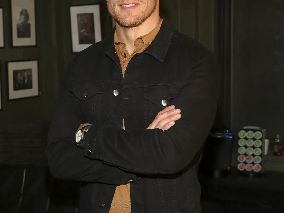 Sam Heughan w czarnej kurtce dżinsowej, z rękami skrzyżowanymi na piersi. W tle zdjęcia i ekspres do kawy.