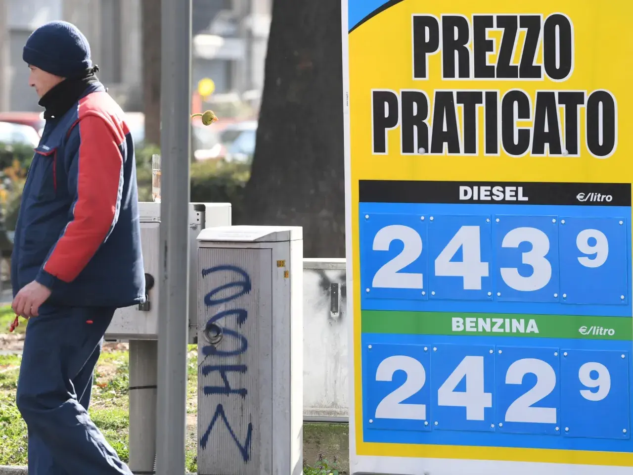 Prezzo praticato per la benzina: 2,429 €/litro. Un uomo in divisa osserva il cartello.