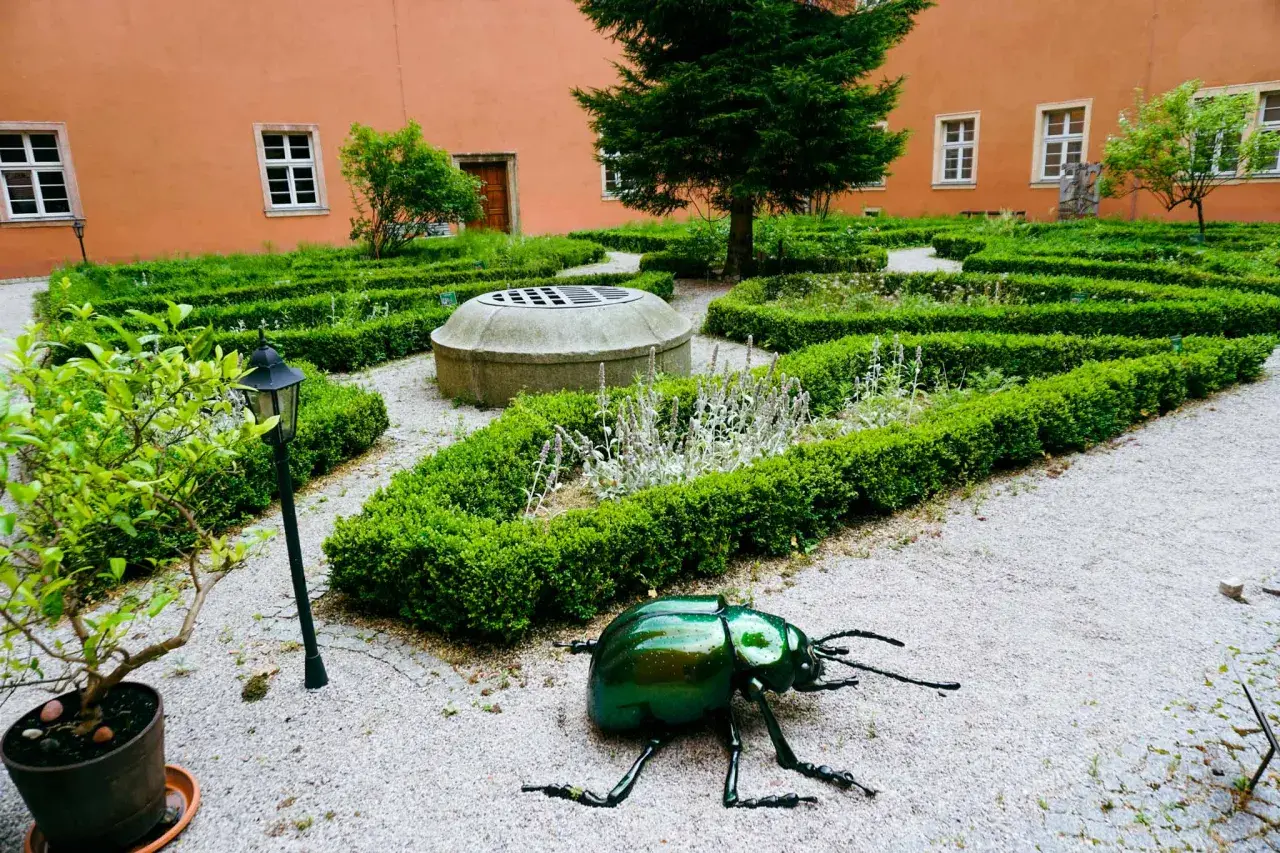 Ogród klasztorny z rzeźbą zielonego chrząszcza, studnią i żywopłotami.