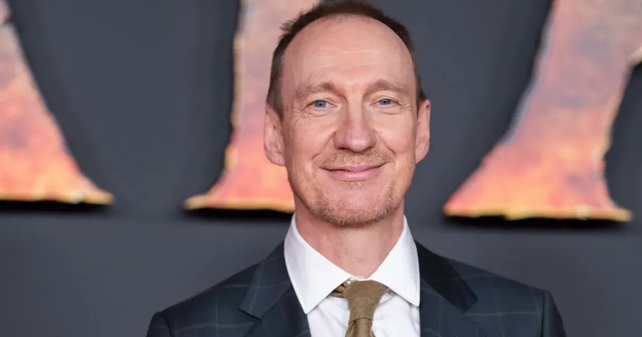 Uśmiechnięty David Thewlis w garniturze i krawacie, z niebieskimi oczami, na tle rozmytych, pomarańczowych kształtów.