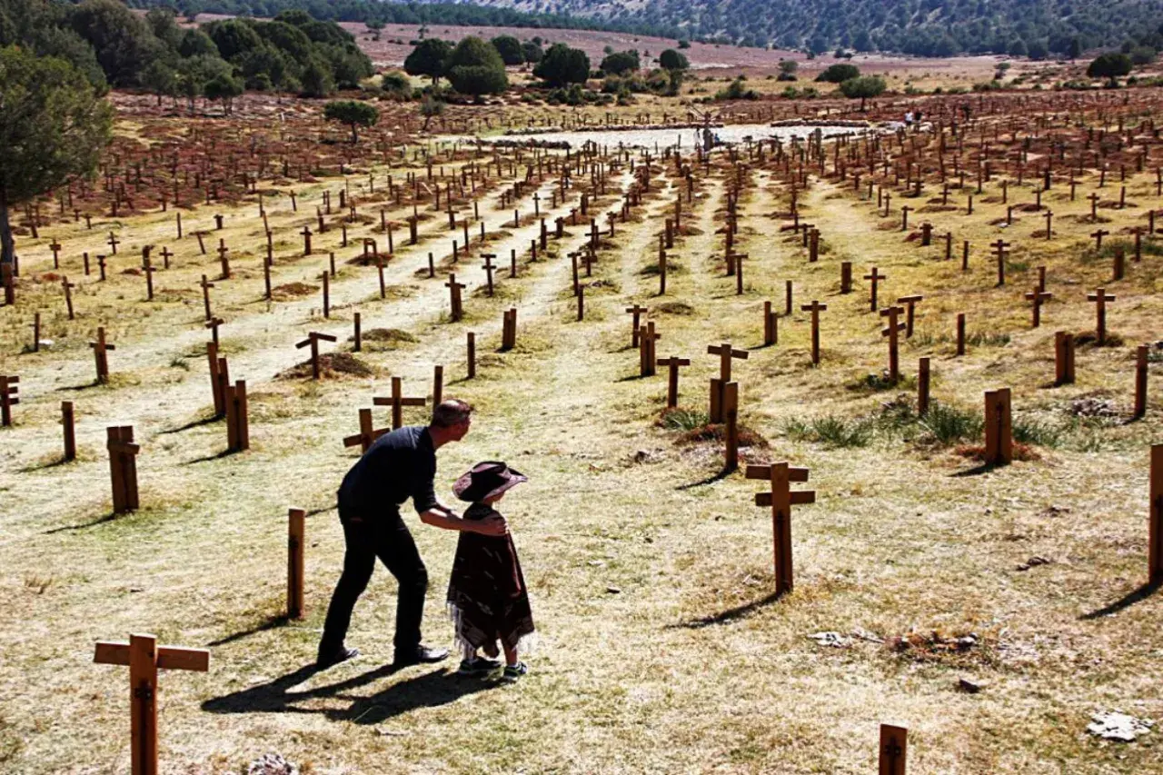 Un hombre y un niño observan un vasto cementerio sad hill, con hileras de cruces de madera bajo un cielo despejado.