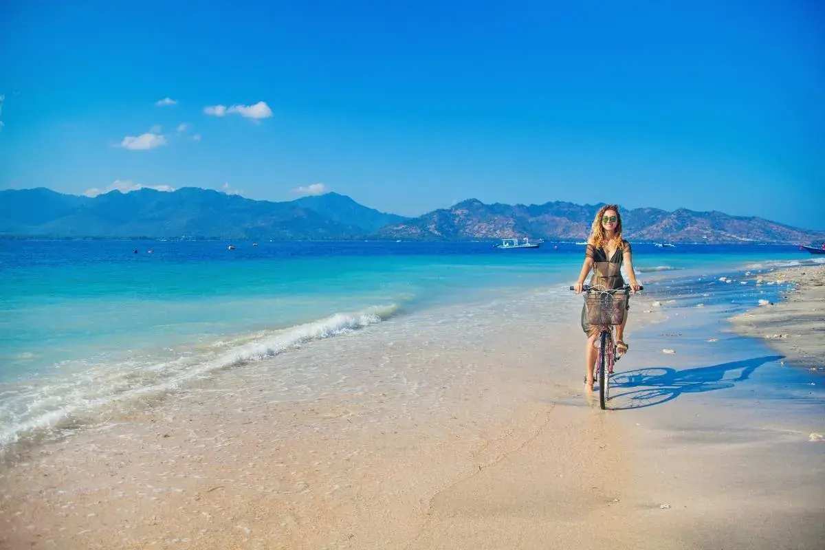 Kobieta na rowerze przemierza piaszczystą plażę z widokiem na turkusowe morze i góry. To idylliczny krajobraz, jak z marzeń o podróży na półwysep Indochiński.
