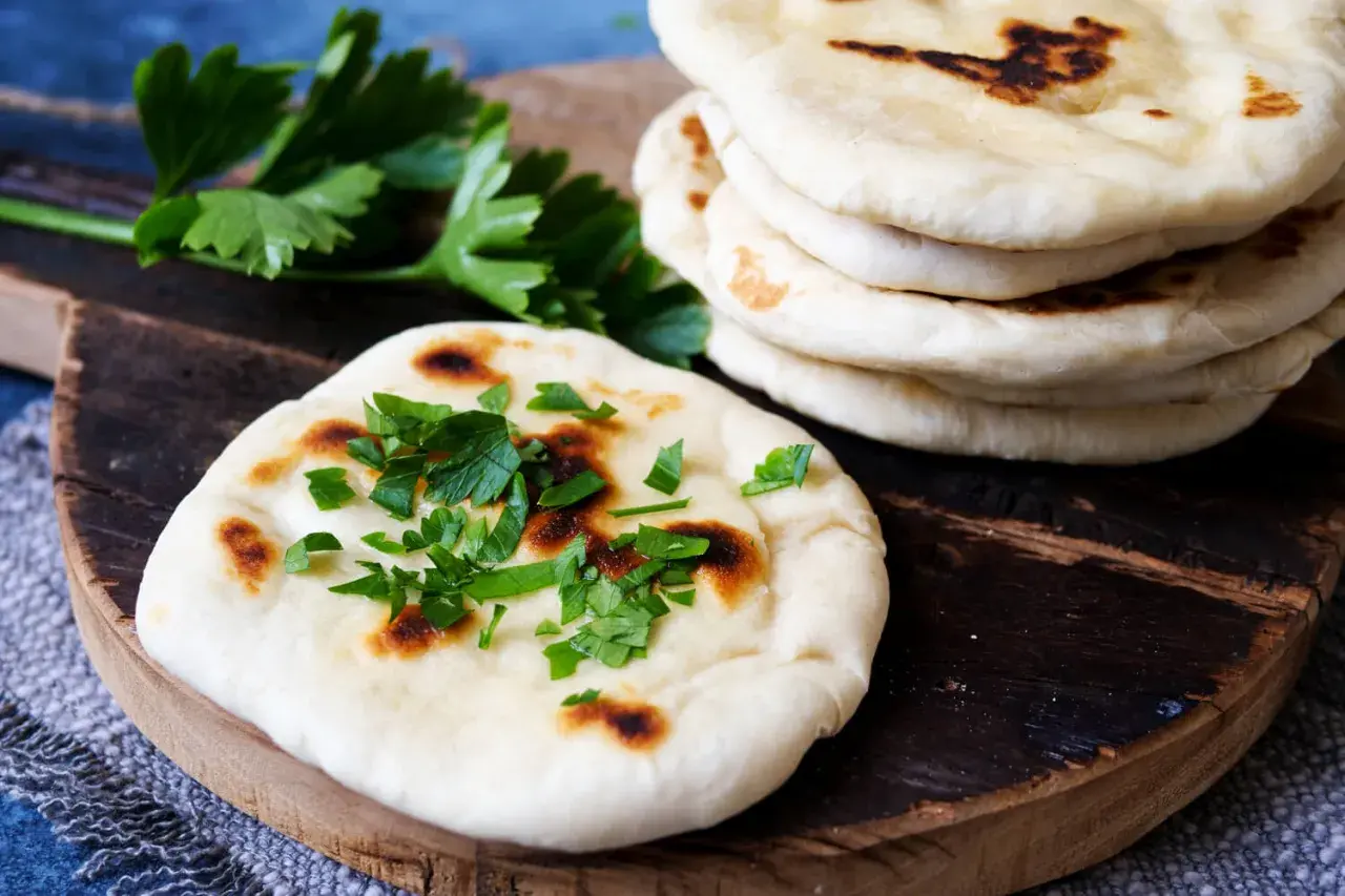 Frisch gebackenes naan brot, bestreut mit Petersilie, liegt auf einem Holzbrett neben einem Stapel weiterer Fladenbrote.