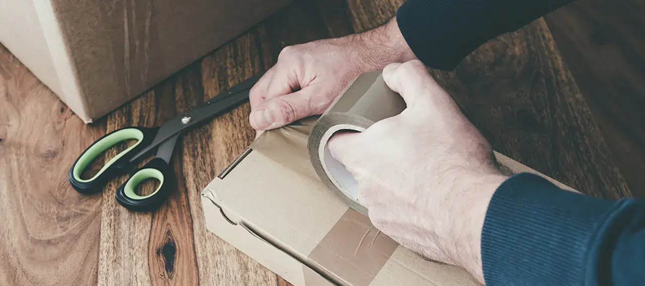 Ręce zaklejają taśmą kartonowe pudełko. Obok leżą nożyczki. Przygotowanie do wysyłki lub obi zwroty.
