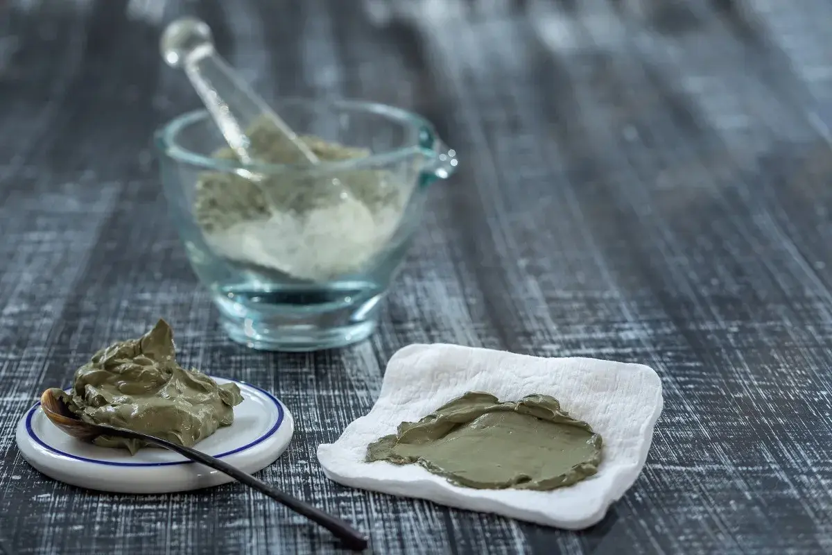 Pâte d'argile verte prête à l'emploi, avec une cuillère sur une petite assiette et une autre portion sur un tissu blanc. Un mortier en verre rempli de poudre d'argile est visible en arrière-plan.