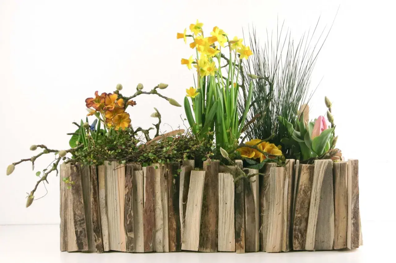 Ein rustikaler Blumenkasten, selbst gebaut aus Holzstücken, gefüllt mit gelben Narzissen, orangefarbenen Primeln und einer rosa Tulpe.