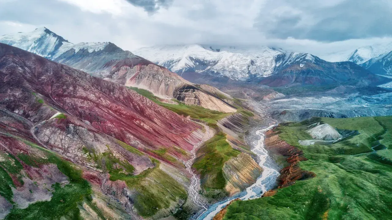 Spektakuläre Kirgisistan Landschaft mit bunten Bergen, einem Fluss und schneebedeckten Gipfeln unter dramatischem Himmel.