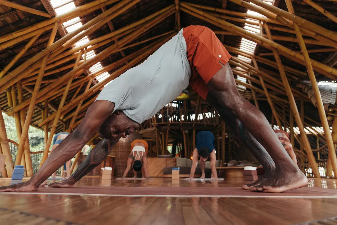 Un homme en posture de chien tête en bas, cherchant à redresser son dos. D'autres pratiquent le yoga dans un studio en bambou.