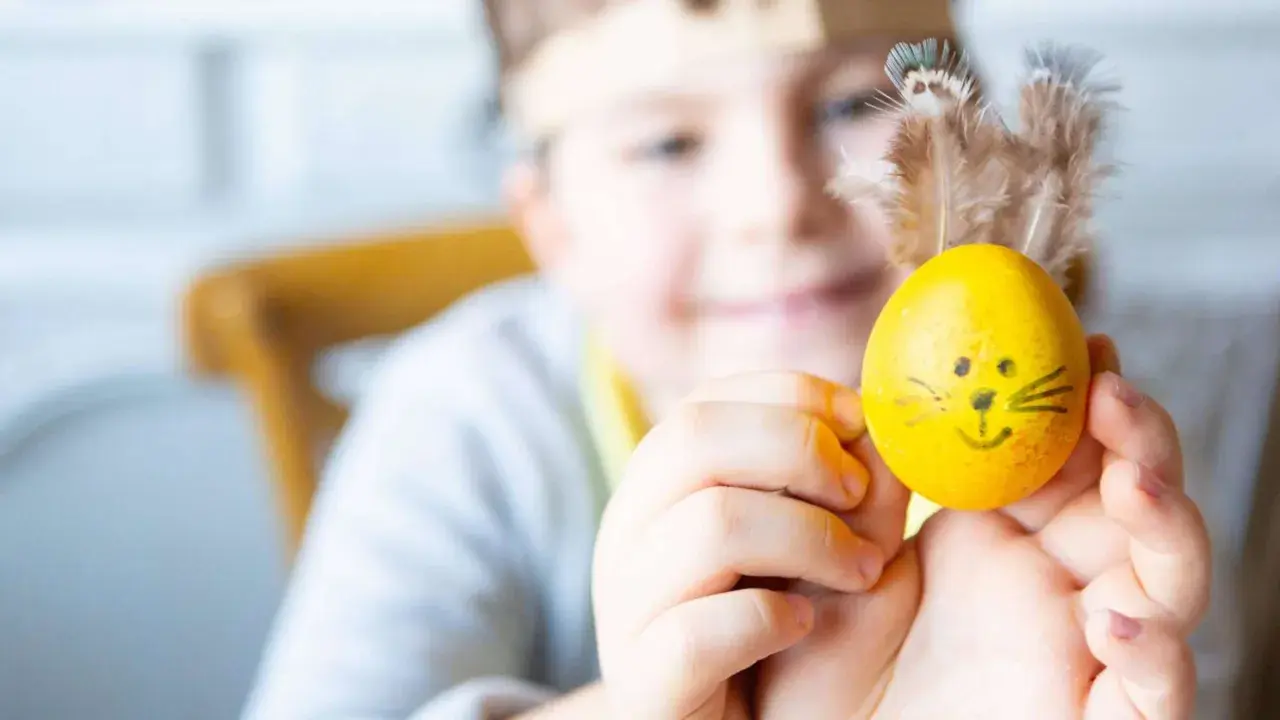 Ein Kind hält ein gelbes Ei mit lustigen Ostermotiv-Gesicht und Federn als Ohren.