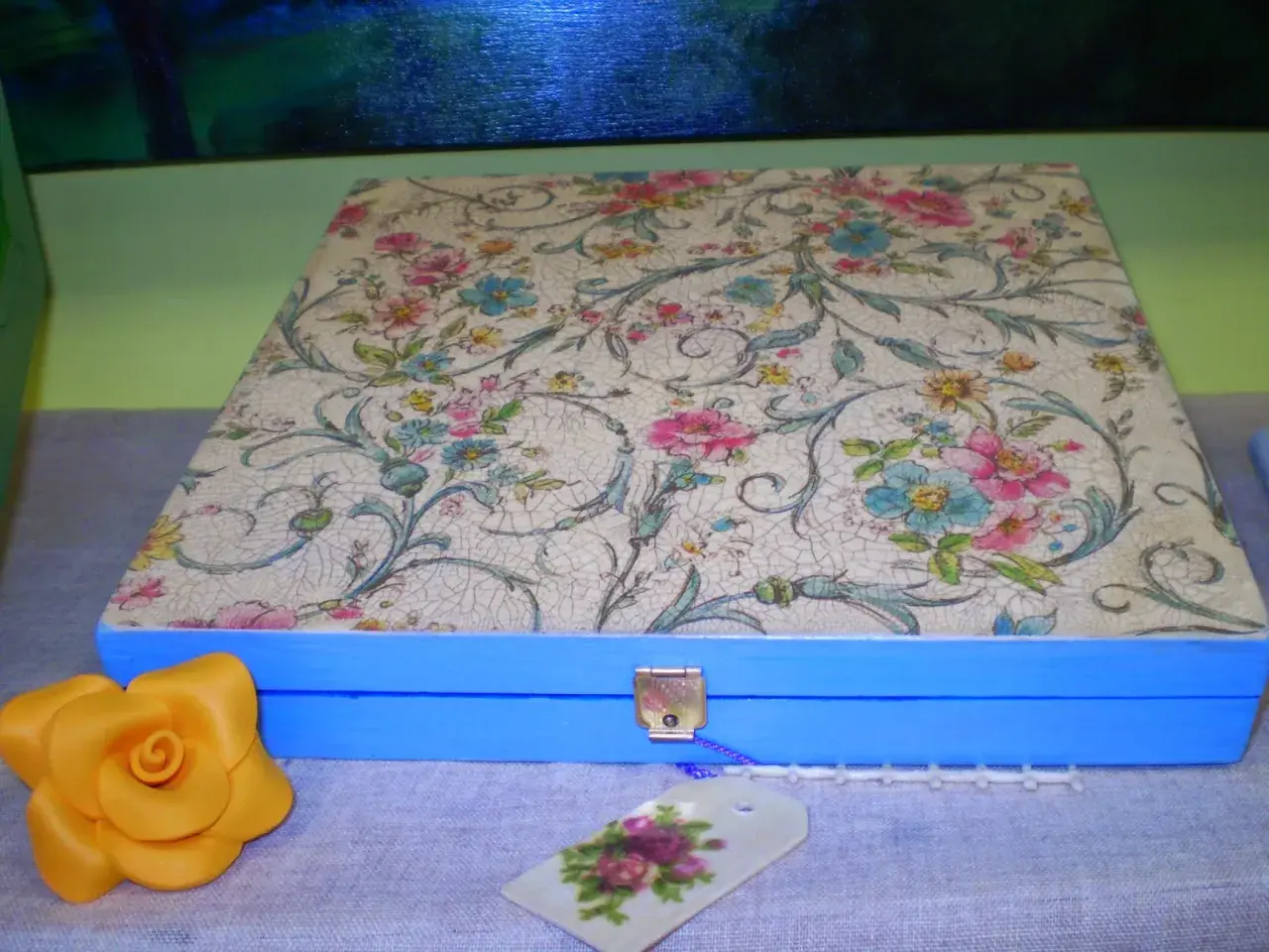Caja azul con tapa decorada con papel decoupage floral. Un detalle de rosa amarilla y etiqueta floral completan la escena.