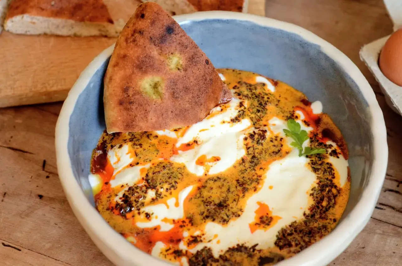Un bol de cilbir recette, avec du pain pita trempé dans une sauce crémeuse et épicée, garni d'herbes fraîches.