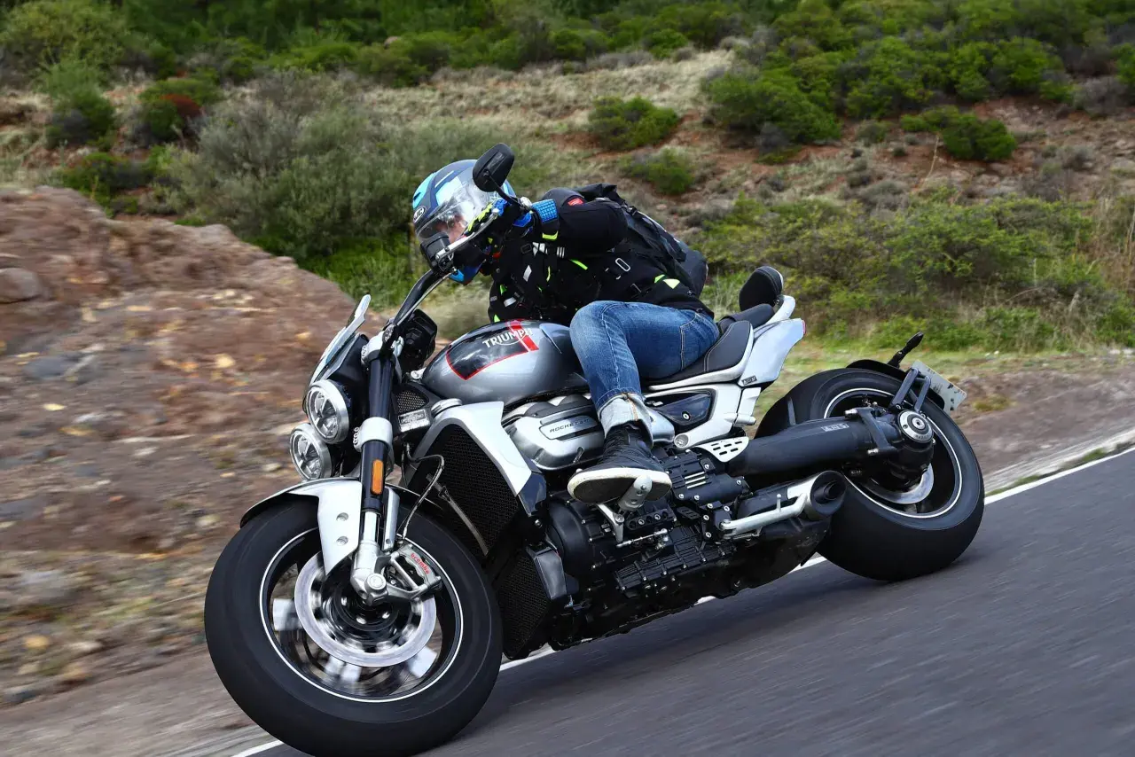 Motocyklista w kasku i kurtce jedzie na motocyklu Triumph Rocket 2 po krętej drodze wśród zieleni.