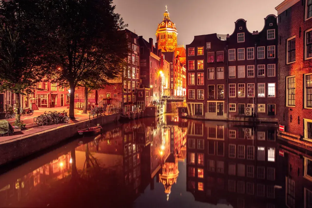 Amsterdam nocą, kanał z odbiciem zabytkowych kamienic i kościoła. Czerwona ulica Amsterdamu rozświetlona latarniami.