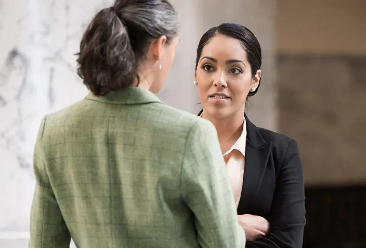 Two women in professional attire converse. One, facing away, wears a green tweed jacket. The other, facing forward, wears a black blazer and light pink blouse, perhaps discussing her northeastern MPA studies.