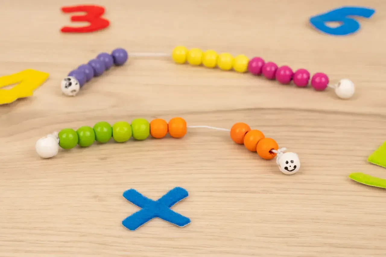 Bunte Perlenketten zum **rechenkette basteln** liegen auf Holz. Zahlen und ein blaues Kreuz sind im Hintergrund.