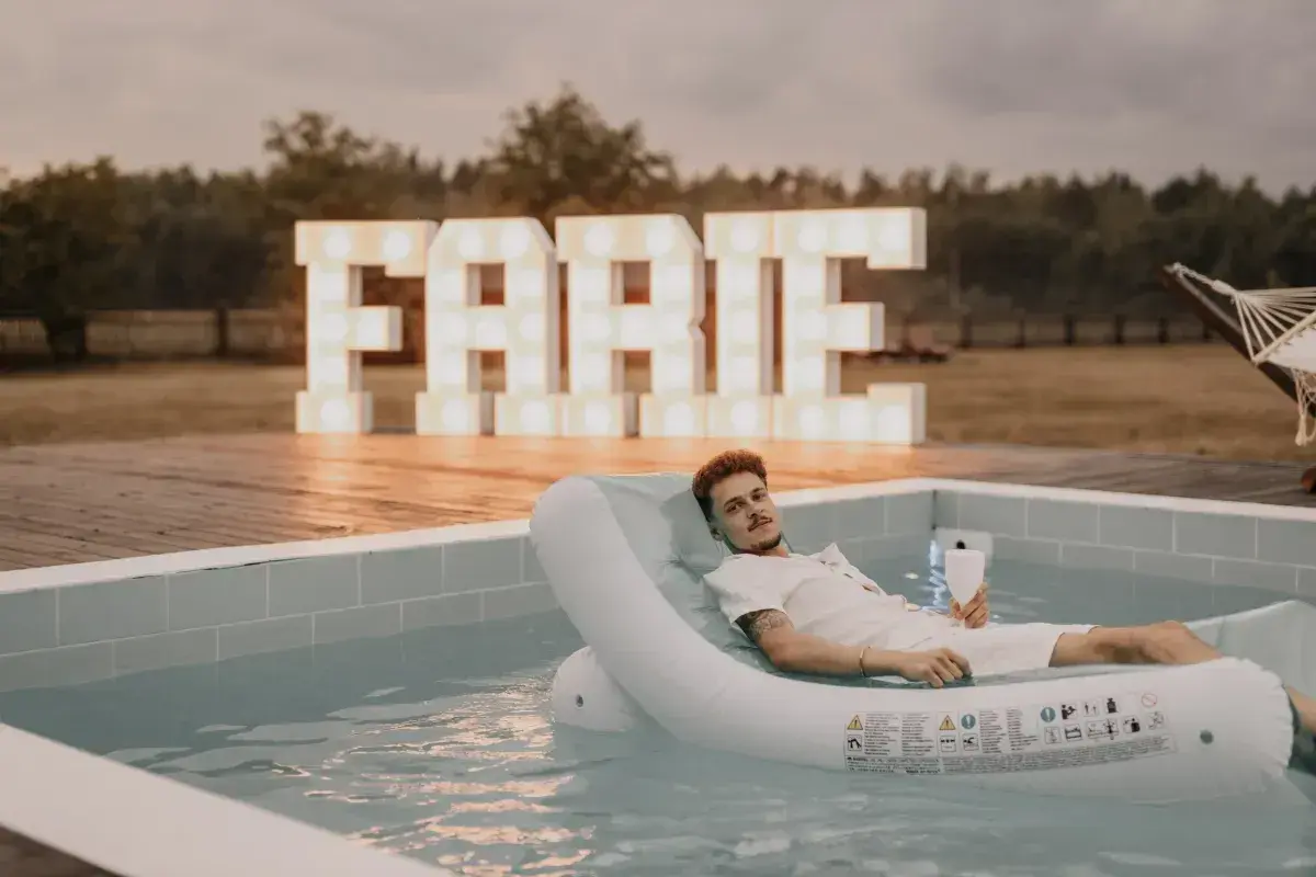 Mężczyzna relaksuje się na dmuchanym materacu w basenie, z wielkimi literami "FARTE" w tle. Idealne miejsce na kawalerski!