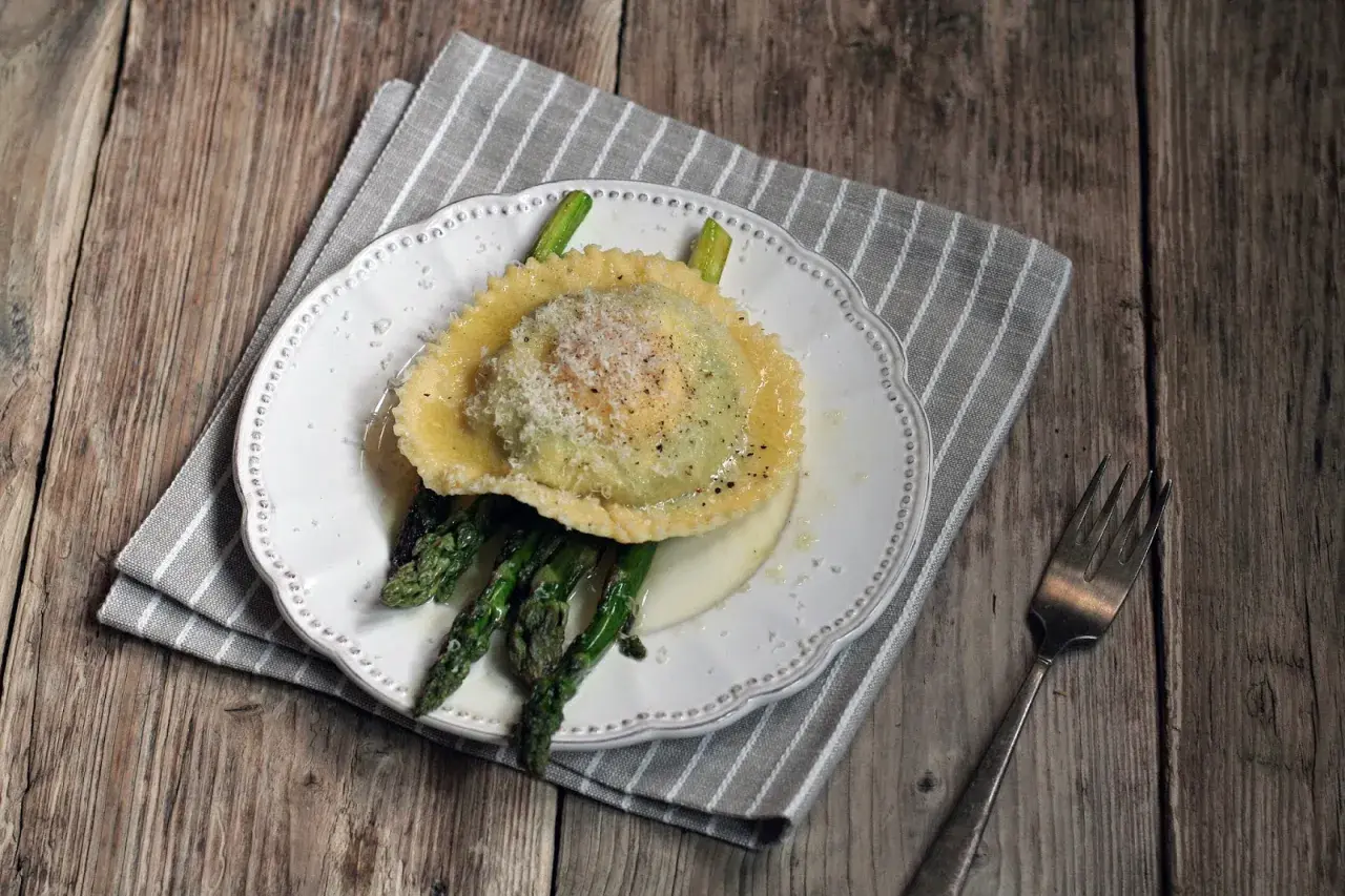 Ravioli z żółtkiem, podane z zielonymi szparagami i posypane parmezanem, na białym talerzu.