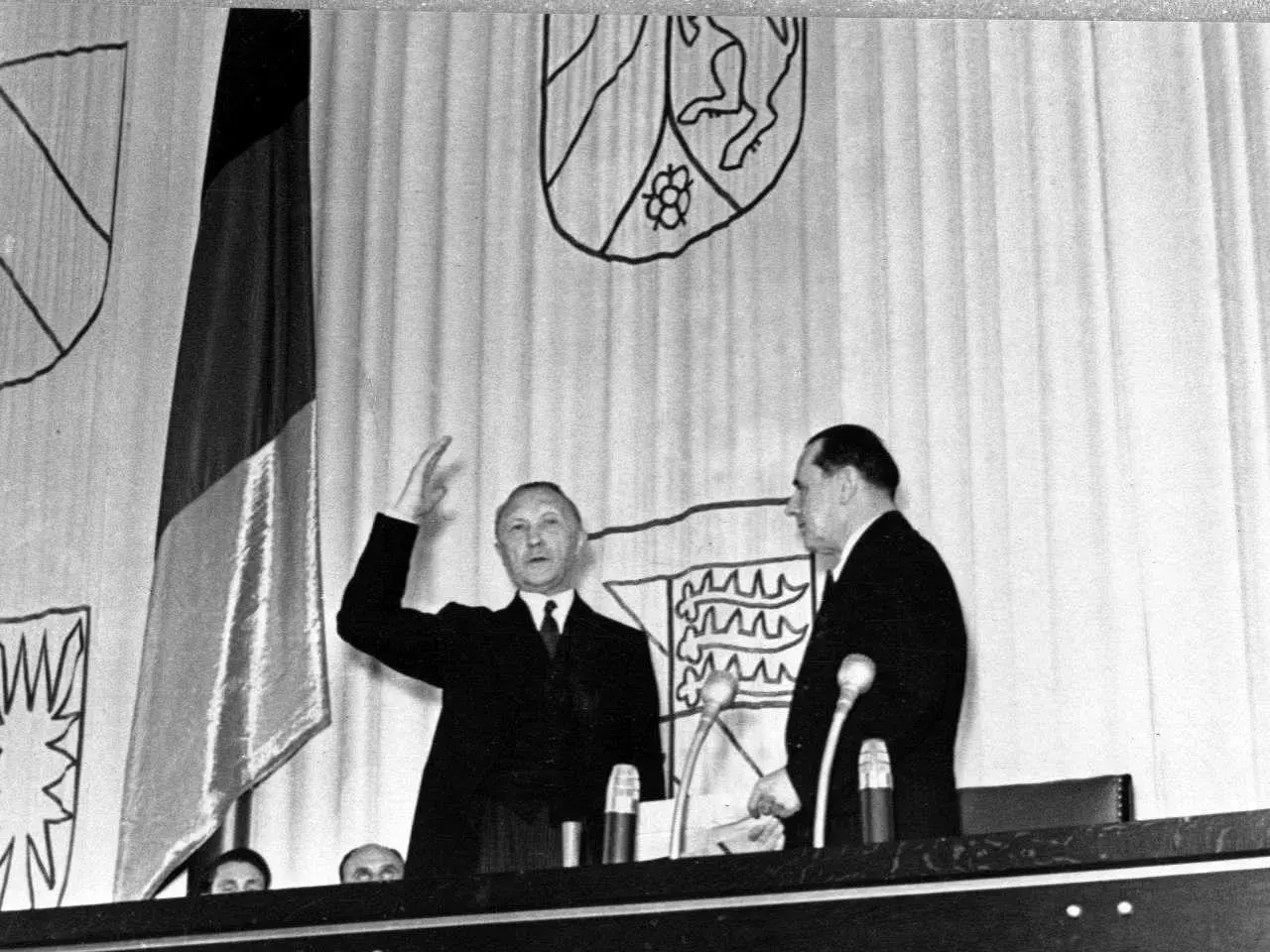 Ein Mann hebt die Hand, neben ihm ein weiterer Mann. Im Hintergrund die deutsche Fahne und Wappen. Der ältere Bundeskanzler spricht.