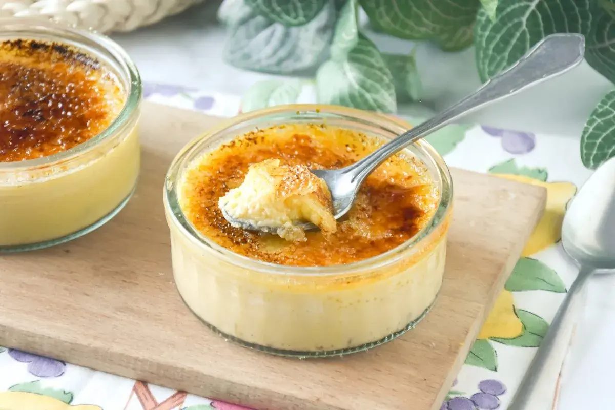 Dos cremosos postres de creme brulee en vasitos de cristal, uno con una cucharada sacada, sobre una tabla de madera.