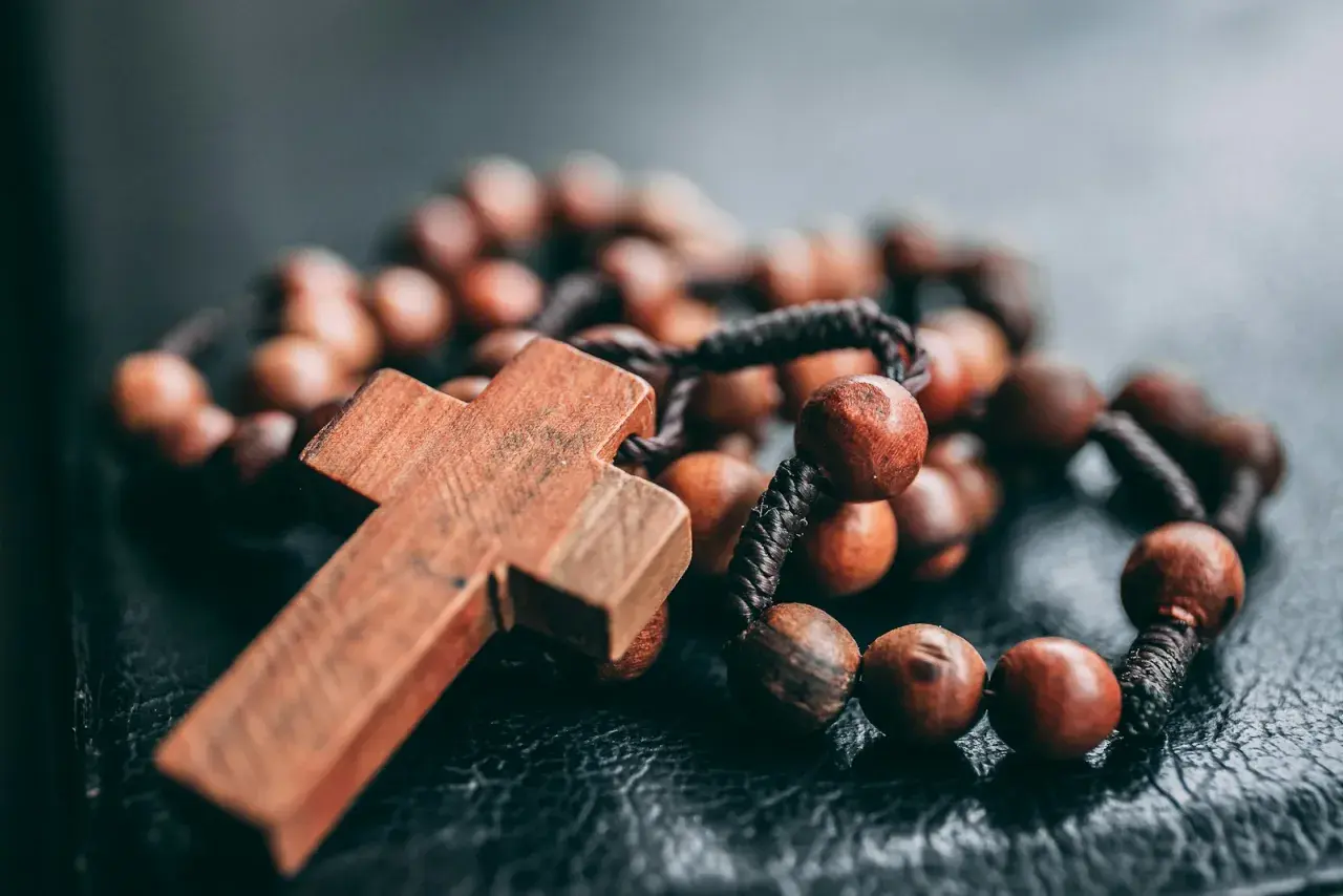 Drewniany krzyż i paciorki różańca, symbolizujące dziesiątkę różańca jako pokutę, leżą na ciemnej powierzchni.