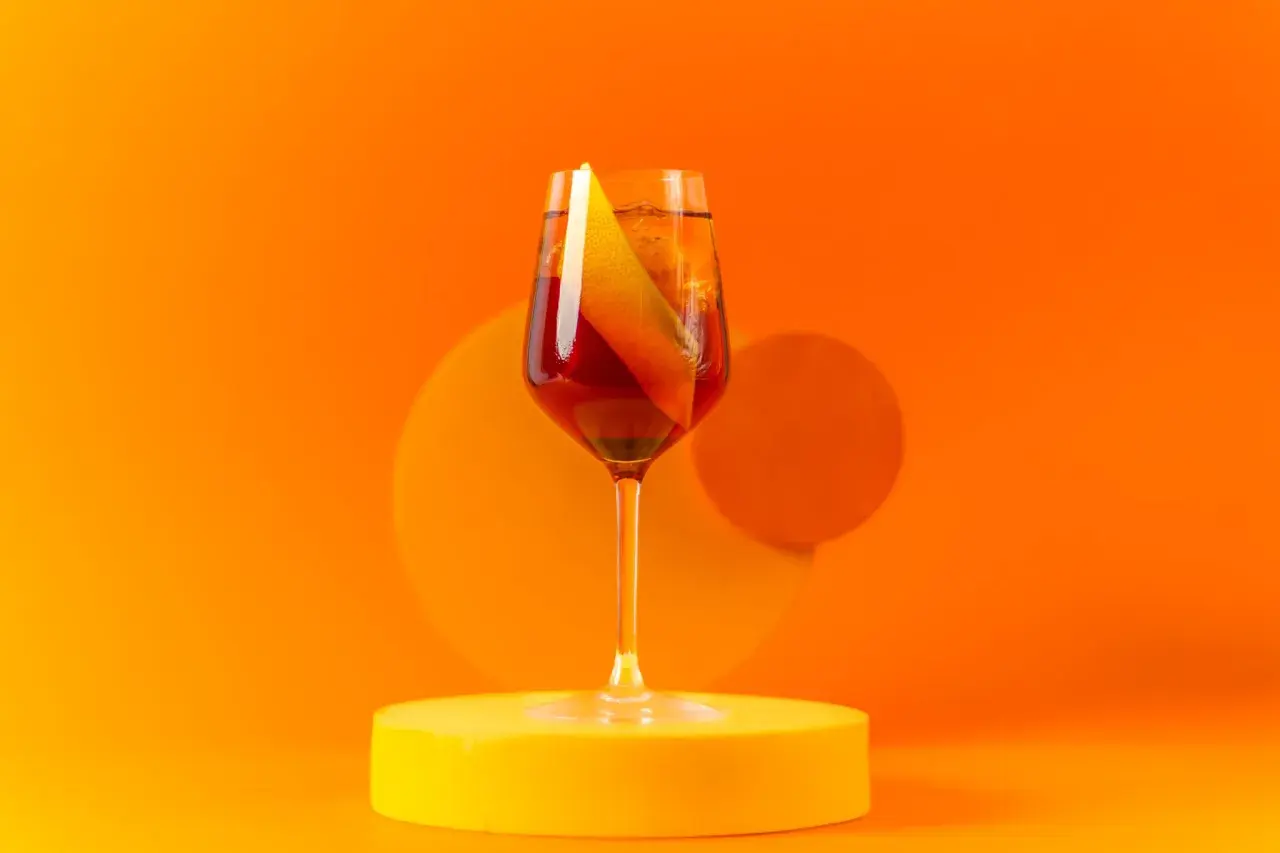 Ein Glas Aperitif mit Orangenscheibe auf gelber Unterlage vor orangefarbenem Hintergrund.