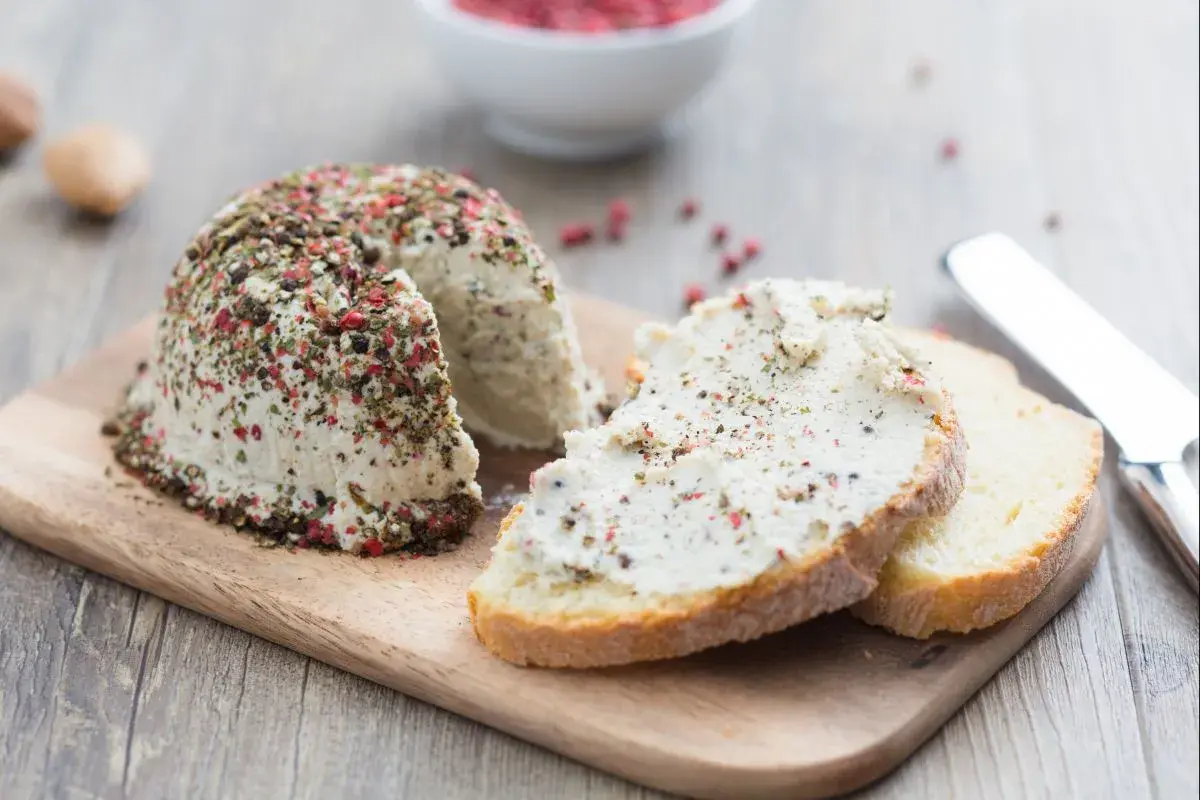 Queso vegano cubierto de pimienta y hierbas, servido en rebanadas de pan rústico.