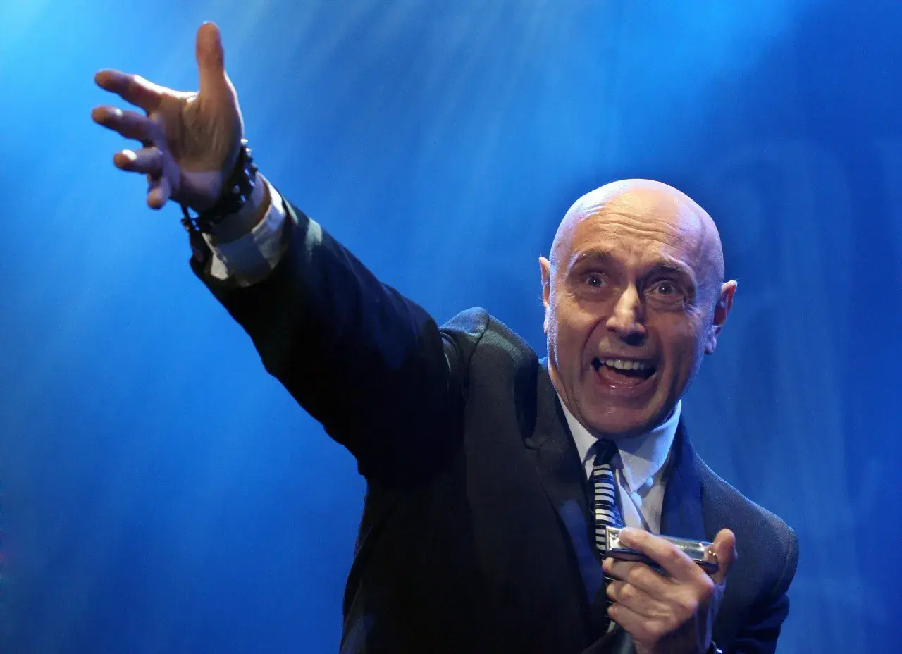 Artysta w garniturze, z podniesioną ręką, gra na harmonijce ustnej. Jego ekspresja sugeruje, że wykonuje energetyczne **shakin dudi utwory**.