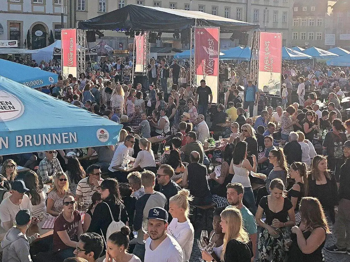 Menschenmenge feiert beim Bamberger Weinfest. Eine Bühne mit Musikern, Fahnen und Zelten. Viele Besucher sitzen an Tischen, trinken und genießen die Atmosphäre.