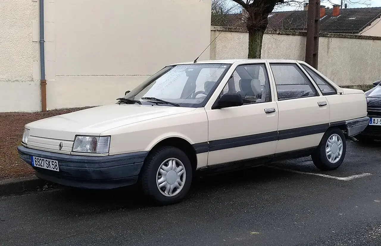 Jasny beżowy sedan Renault 21 z lat 80. stoi na mokrej drodze.