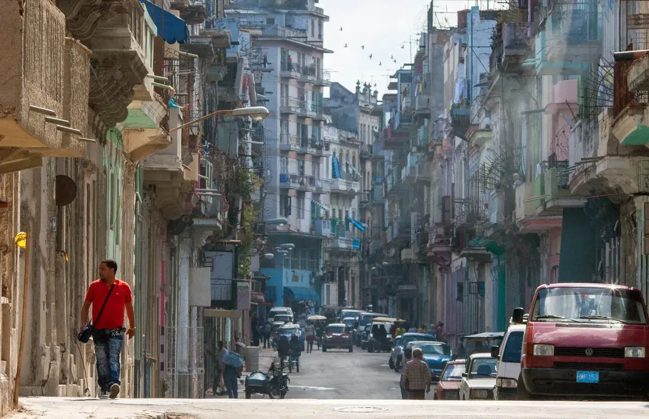 Photographie de rue d'une rue cubaine animée, avec des bâtiments colorés et délabrés, des voitures anciennes et un homme marchant seul.