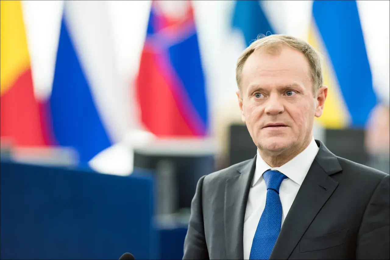 Donald Tusk w garniturze i niebieskim krawacie na tle flag.