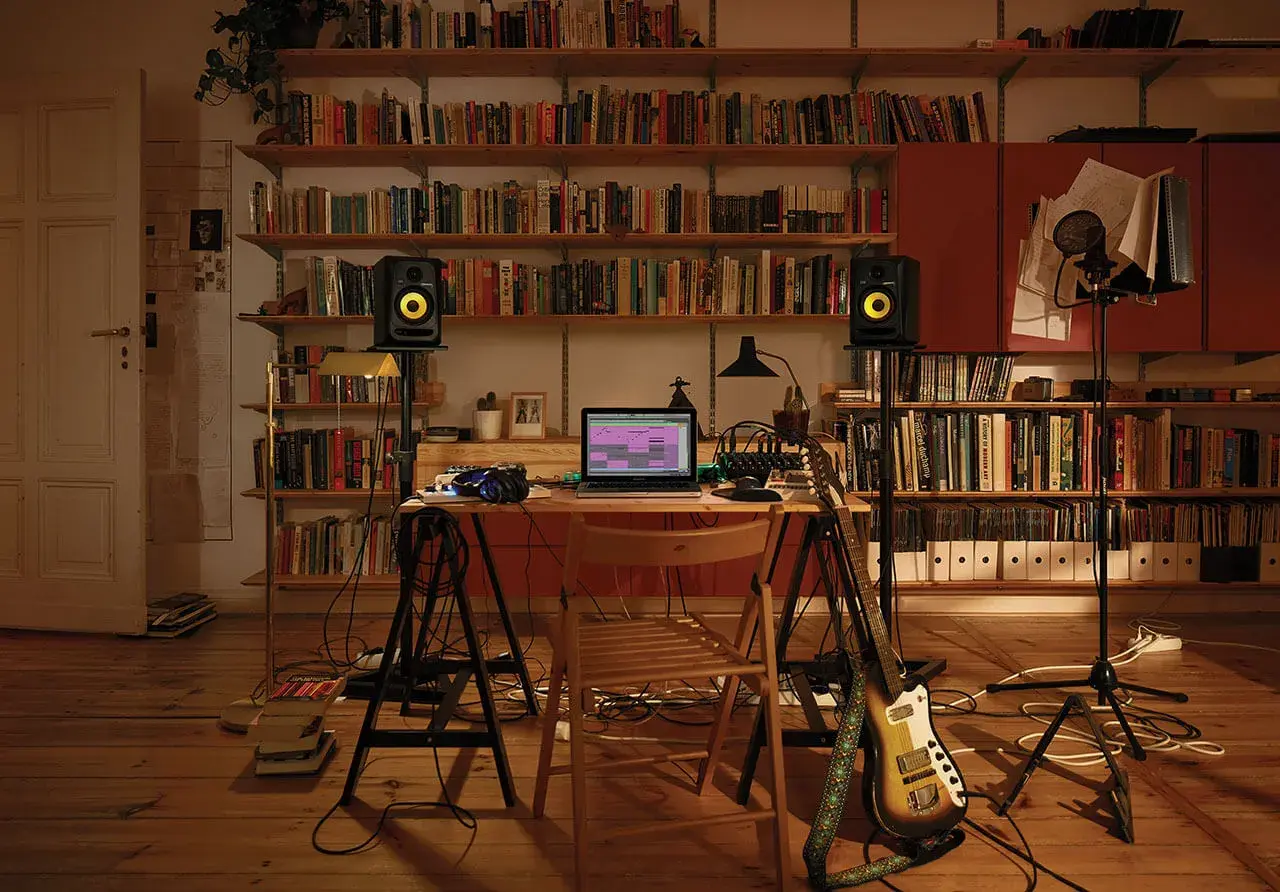 Bureau de musique avec ordinateur affichant un logiciel comme FL Studio, enceintes, guitare et microphones, entouré d'étagères remplies de livres.