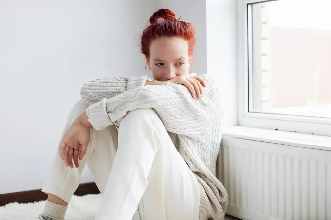 Ragazza con capelli rossi seduta vicino a una finestra, avvolta in un maglione bianco. Potrebbe riflettere su come l'olanzapina a cosa serve per affrontare momenti difficili.